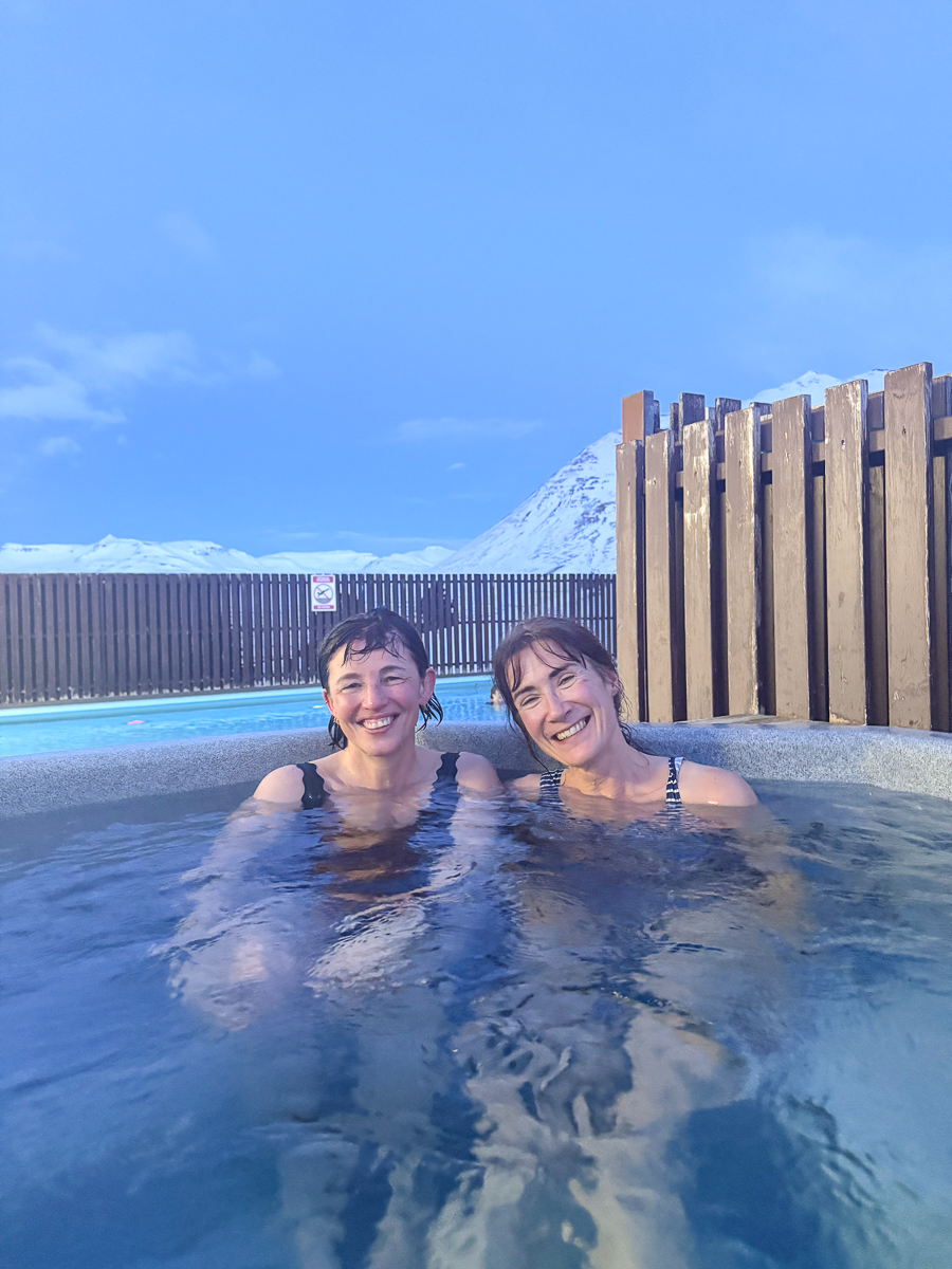 Women smiling in a hot tub during winter geothermal bathing, with snowy mountains in North Iceland