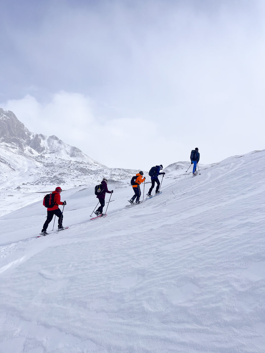 Snowshoe hikers trek across a snowy mountain slope under a bright winter sky