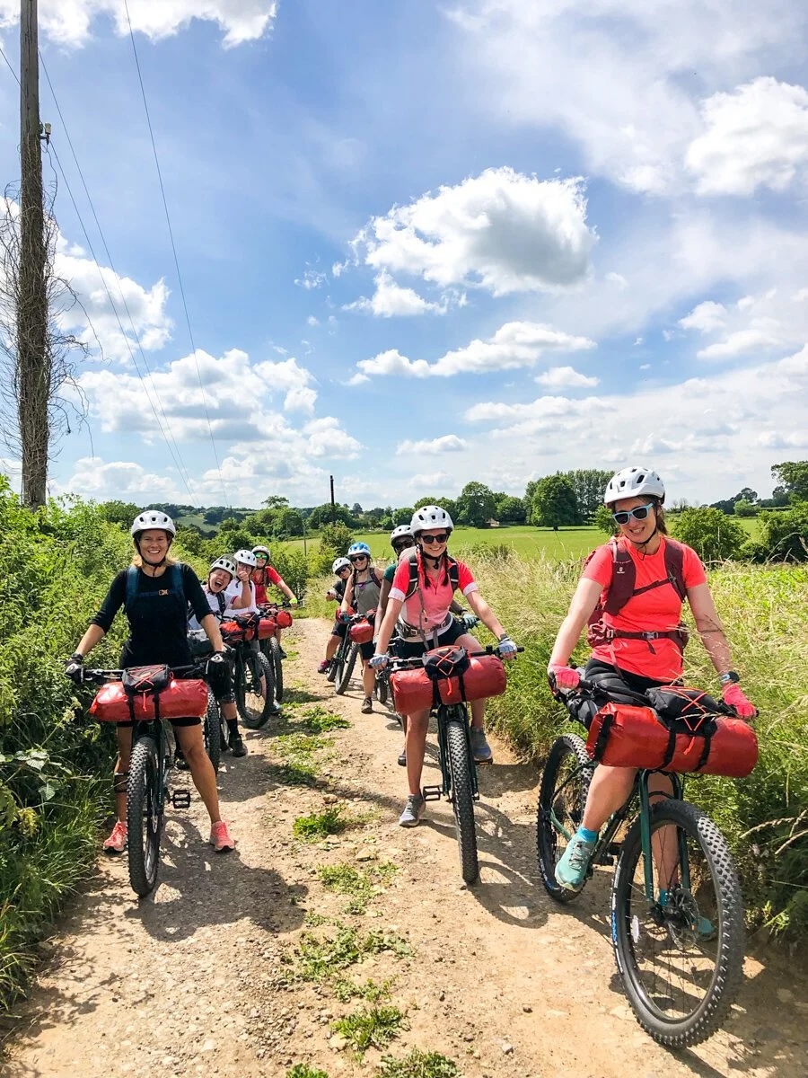 Women bikepacking on gravel path through green countryside, with helmets and pannier bags