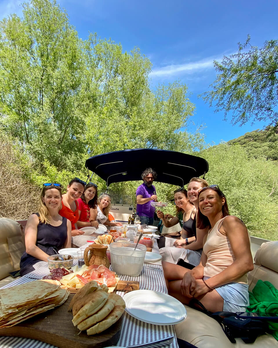 Women enjoying a picnic outdoors on a terrace with an open awning, surrounded by green hills and blue sky