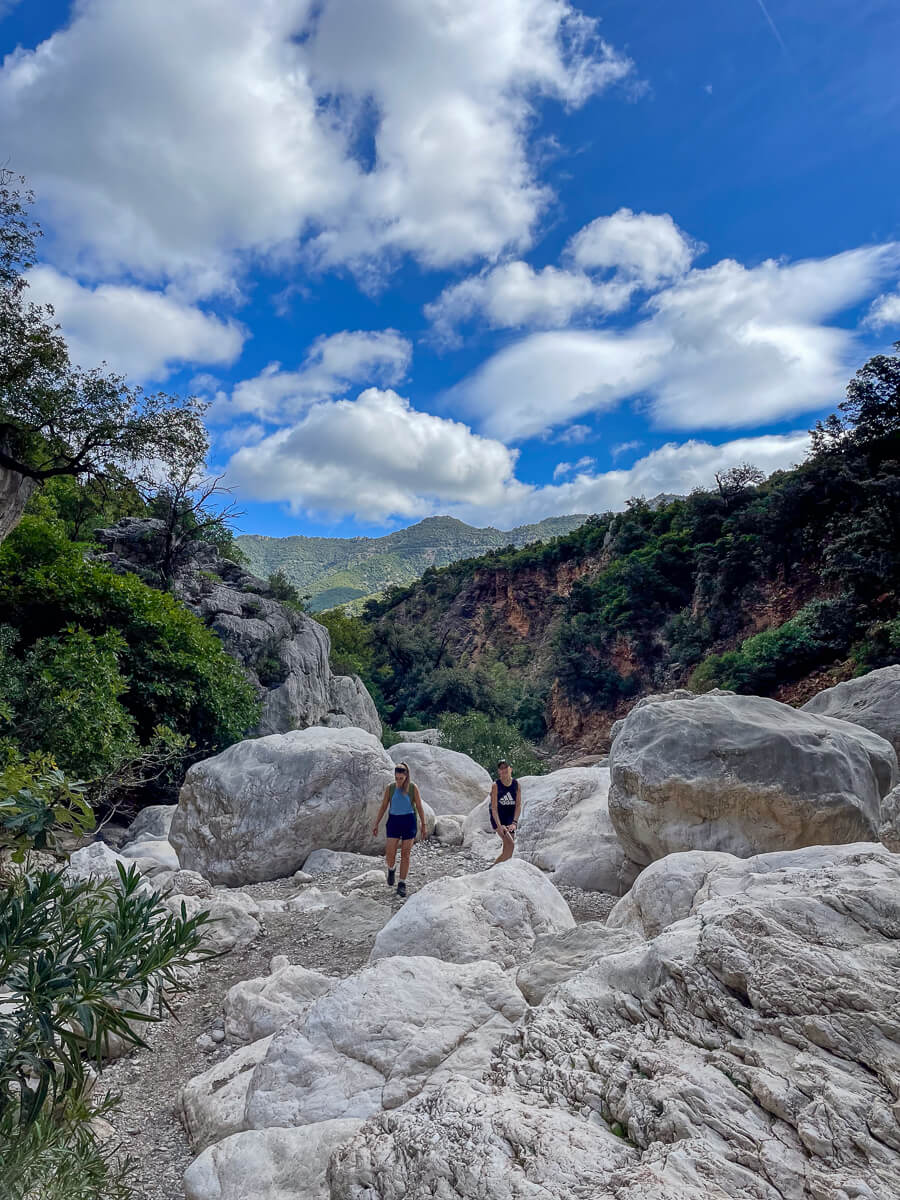Women hiking on a rocky trail in East Sardinia mountains under a blue sky with scattered clouds