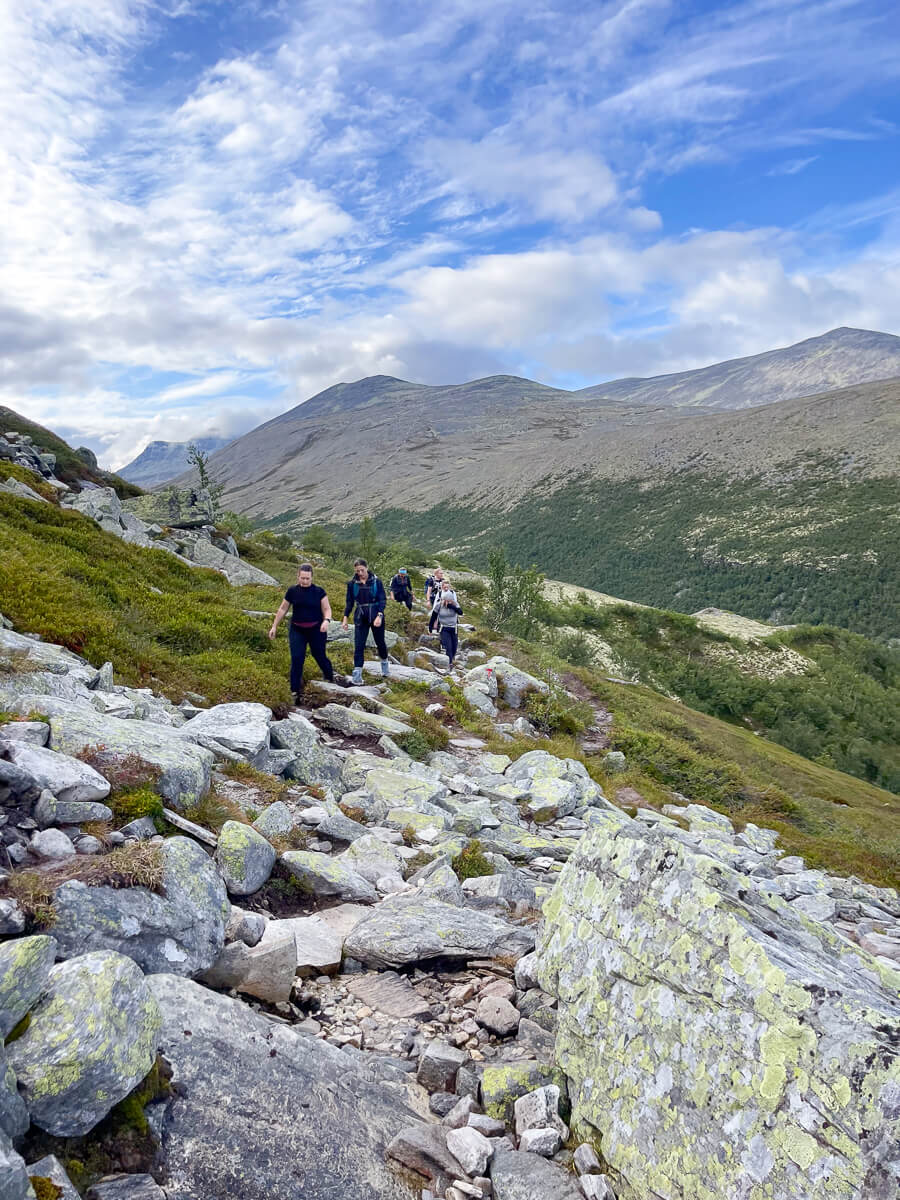 Women hiking on a rocky mountain trail with peaks and valley views under a blue sky
