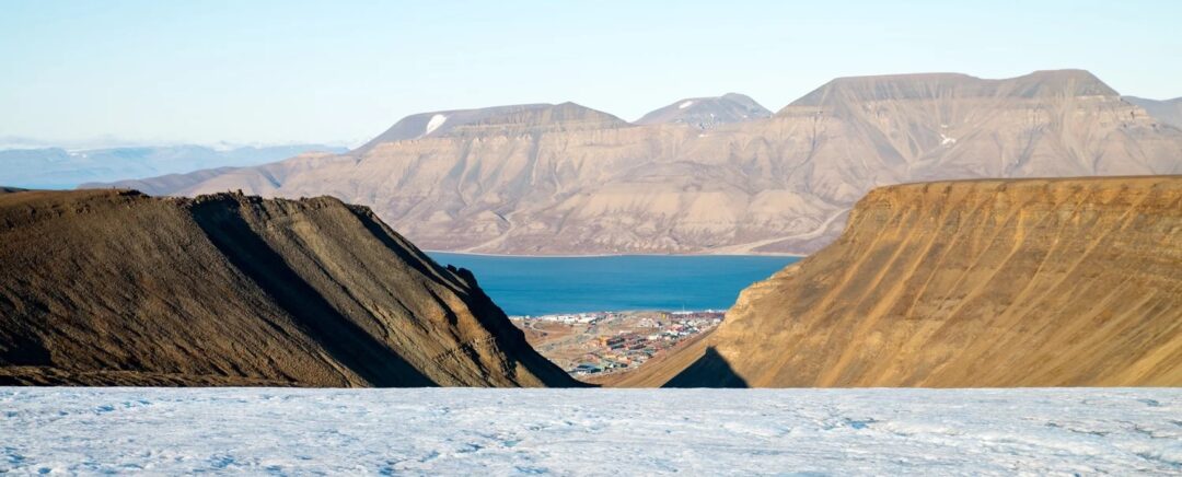 Arctic wilderness landscape in Svalbard with steep mountains, icy water and a distant settlement by the fjord