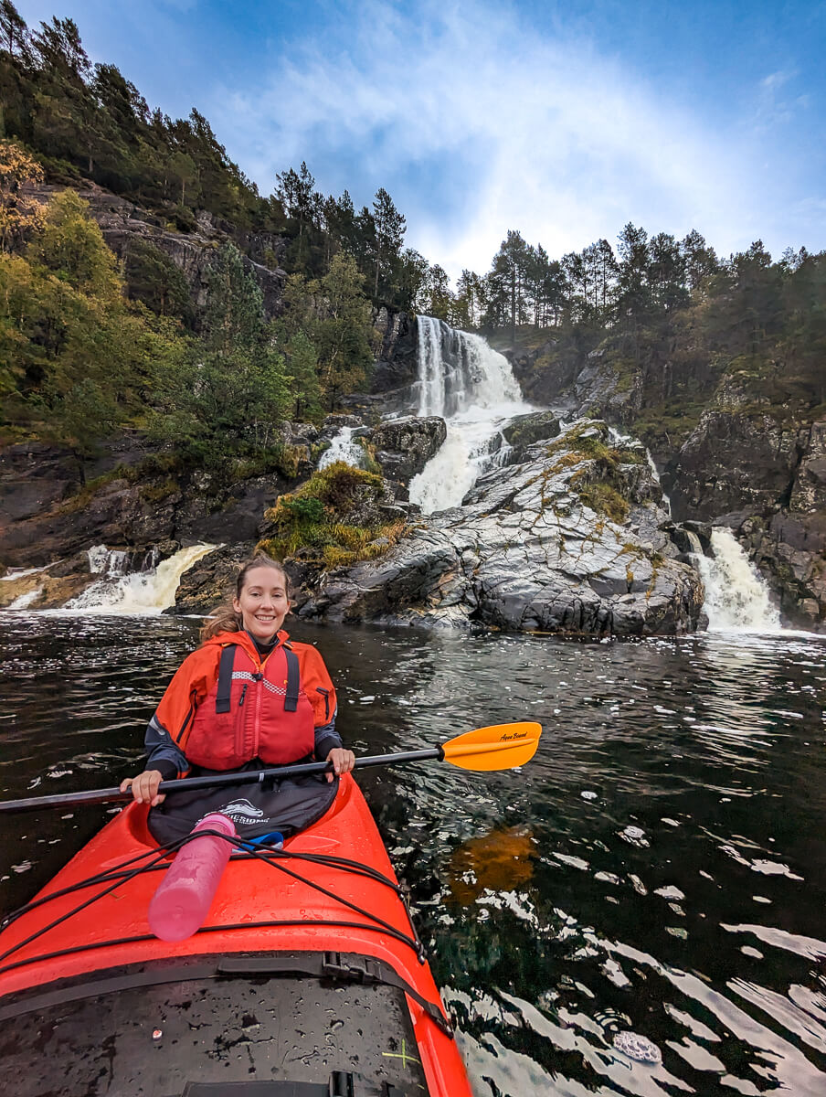 Woman kayaking past a waterfall in a rocky, forested mountain landscape