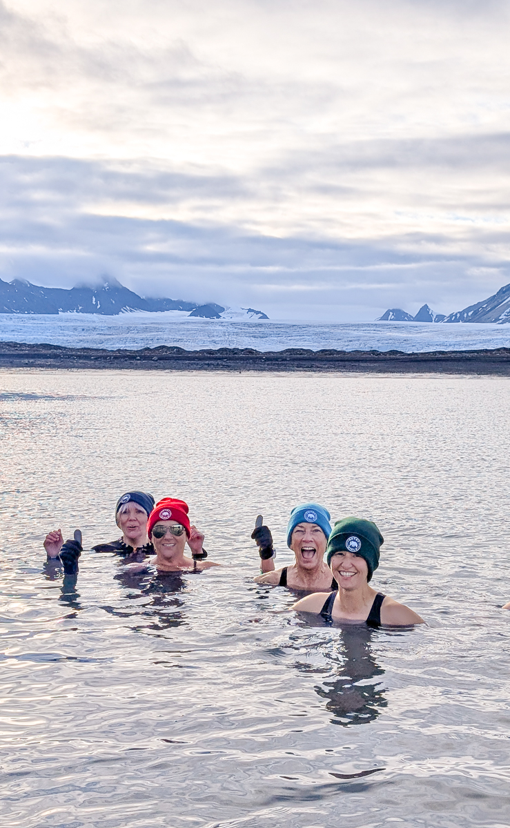 Women in wetsuits cheer while swimming in icy waters on a Svalbard Arctic expedition with glaciers behind