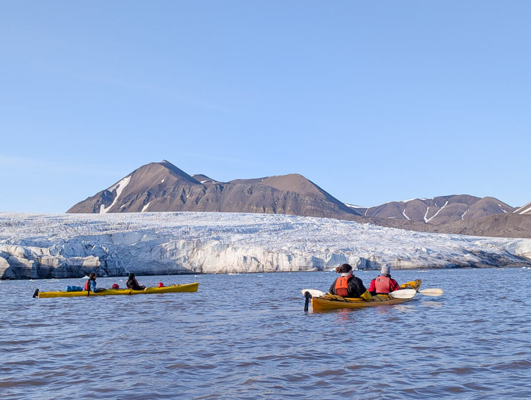 Kayakers paddle on calm water with a glacier and mountains in Svalbard, Arctic wilderness summer expedition.