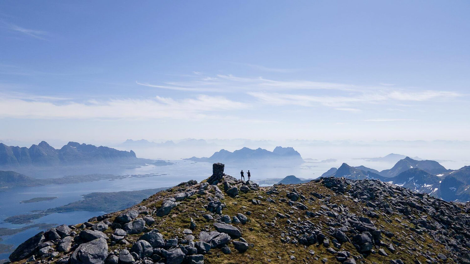 Scenic coastal mountain viewpoint from a rocky ridge, with fjord and islands below under a misty sky