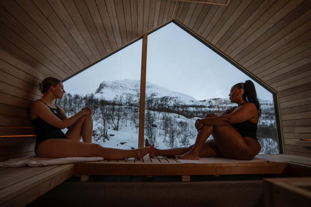 Two women relaxing in a wooden sauna, with snowy mountain views through the window