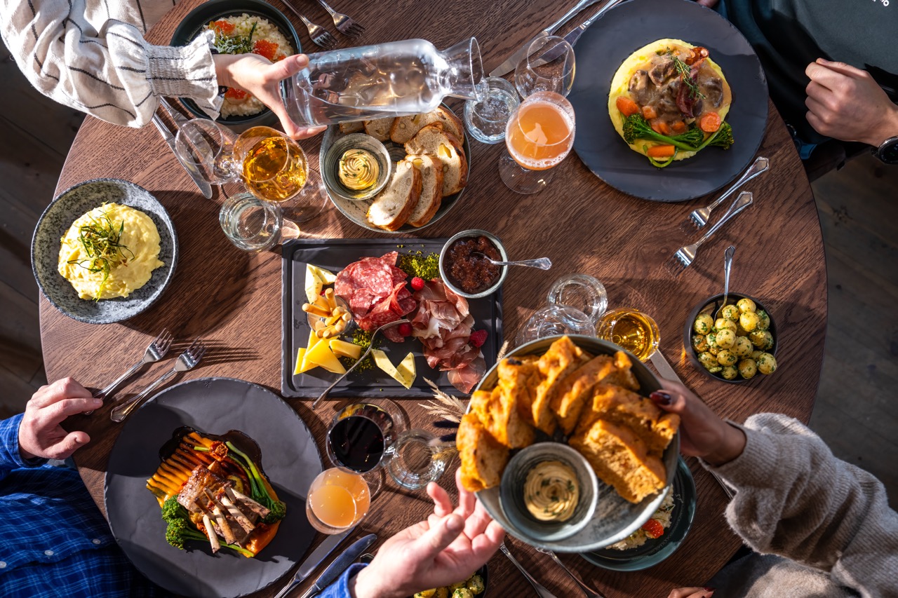 Diners sharing a meal and drinks around a wooden table with bread, pasta, salad and olives