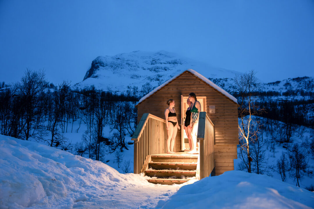 Two women step outside a wooden cabin in snowy mountains at dusk under a clear blue sky