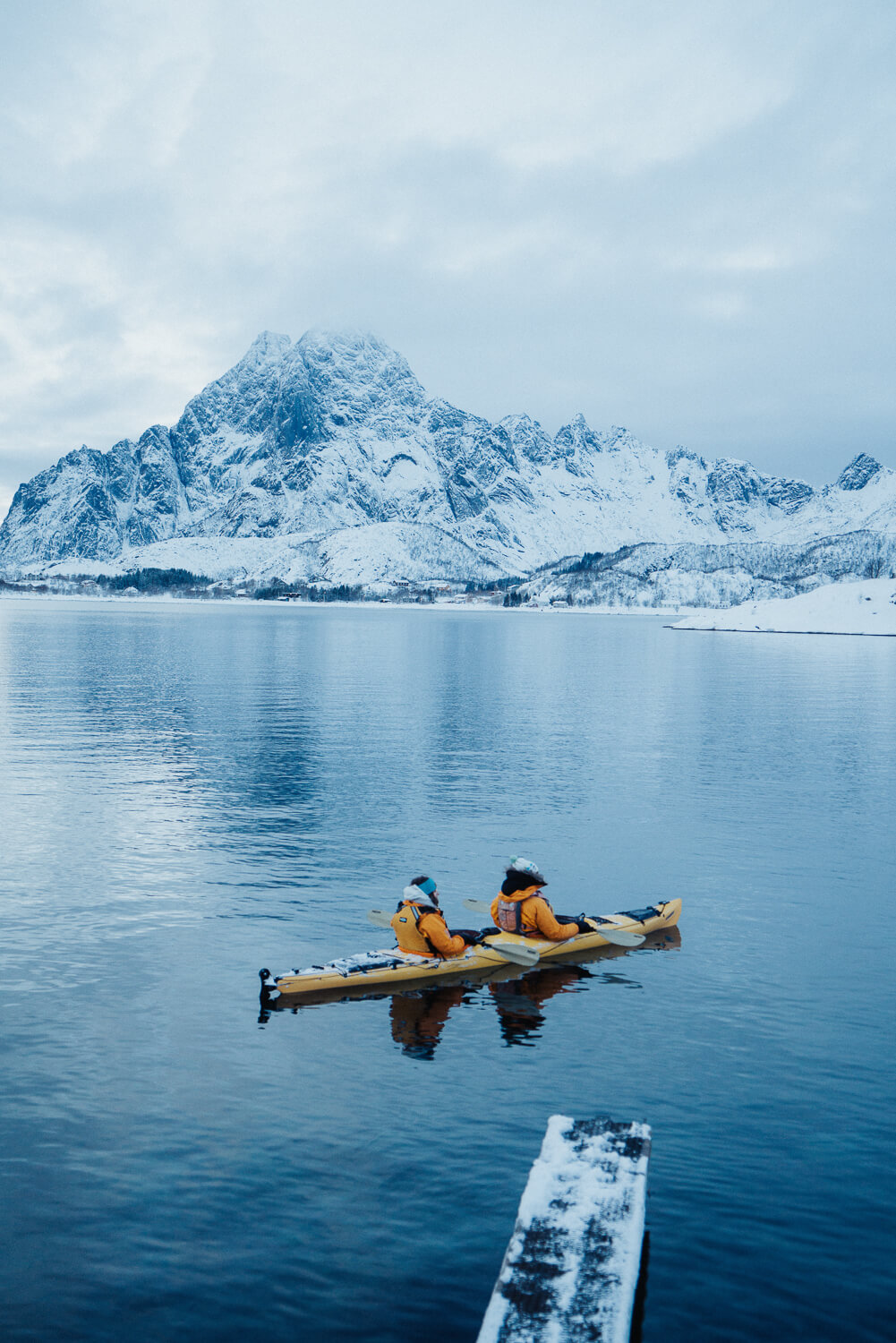 Kayakers on a still fjord in Lofoten with snow-capped mountains in the background.