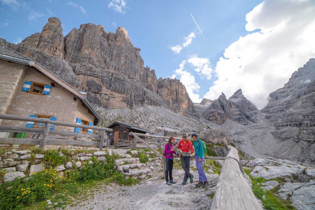 Women hiking on a trail with the Dolomites mountain range in Italy, with a mountain hut in view.