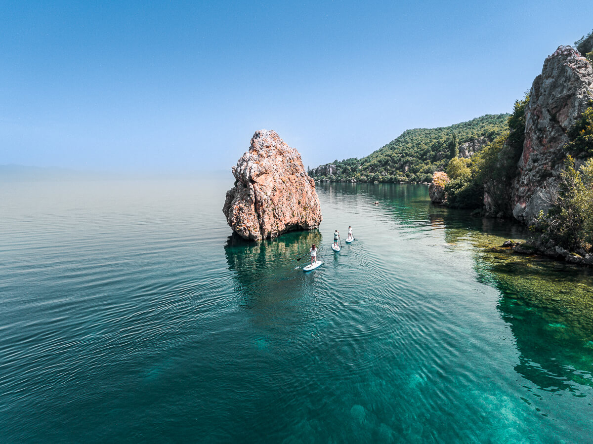 Women stand-up paddleboarding on Lake Ohrid, North Macedonia, beside rocky cliffs.