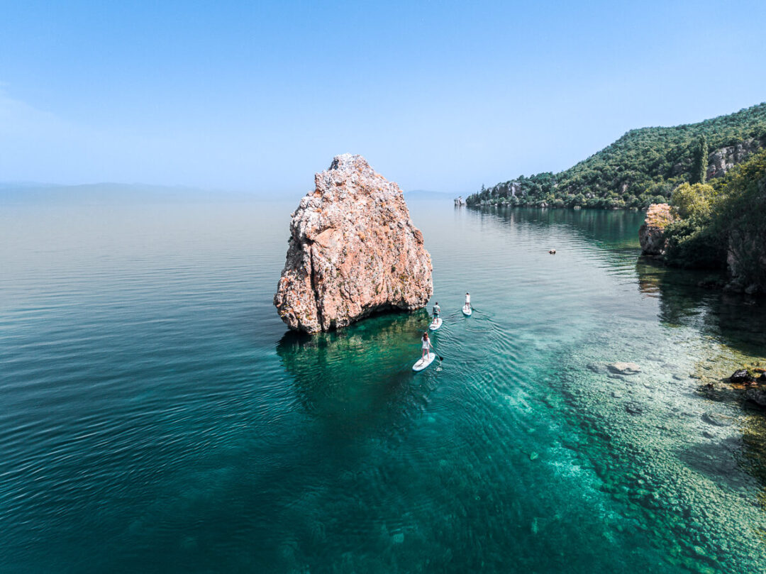 Paddleboarders on turquoise water near a rocky outcrop at Lake Ohrid, North Macedonia.