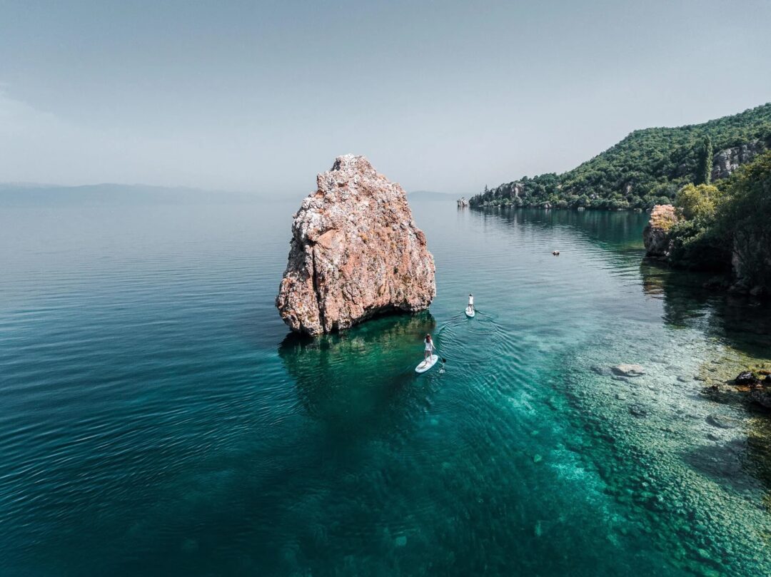 Isolated rock in clear blue lake water with boats on a tranquil shoreline