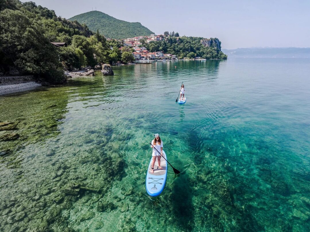 Women paddleboarding on a calm lake near Ohrid, North Macedonia, with forested shoreline and hills in view