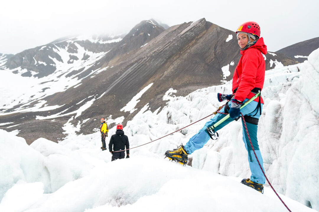 Woman ice climbing on a Svalbard glacier, in bright red jacket and helmet with rope in snowy mountains