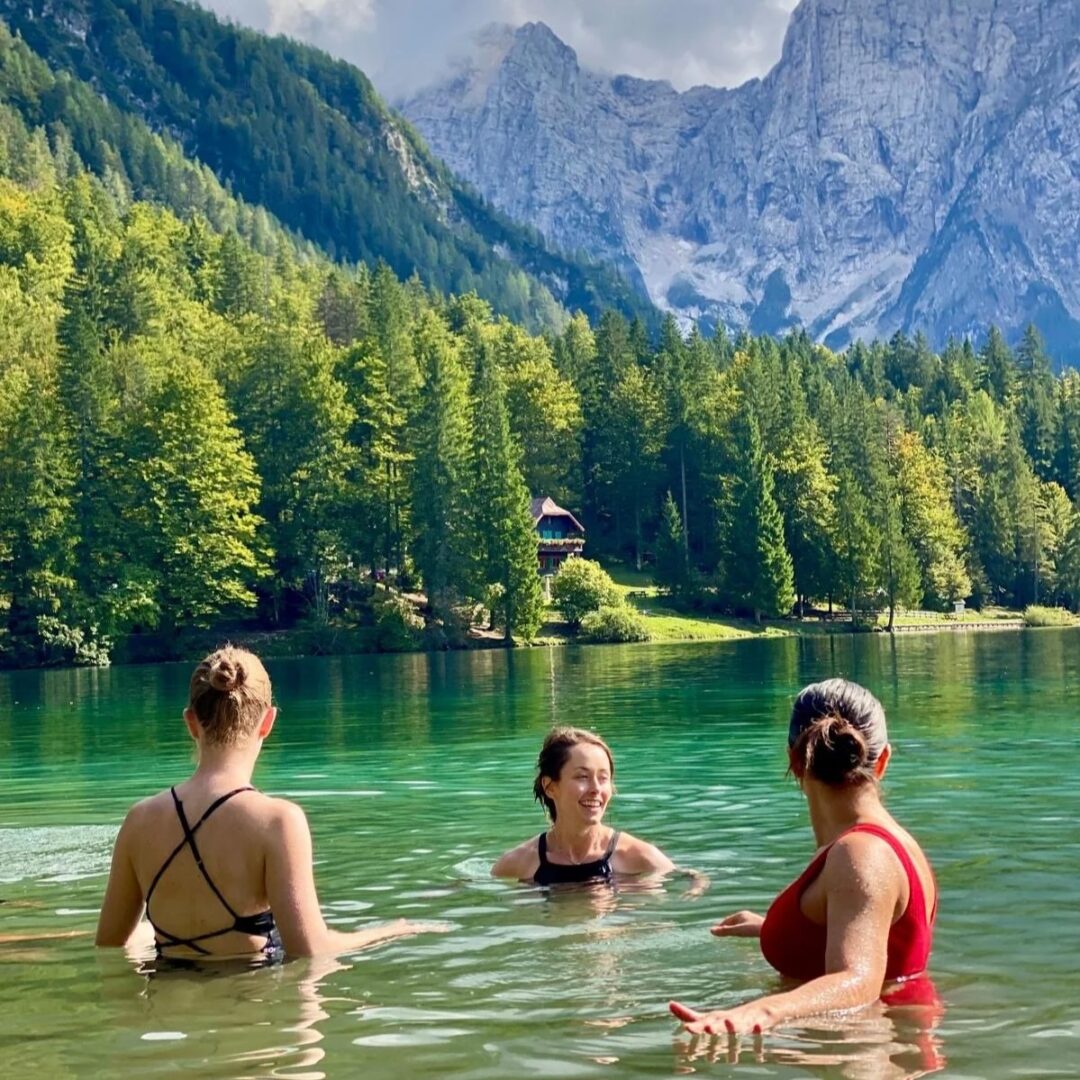 Women swimming in a calm mountain lake with forested slopes and snowy peaks in the background