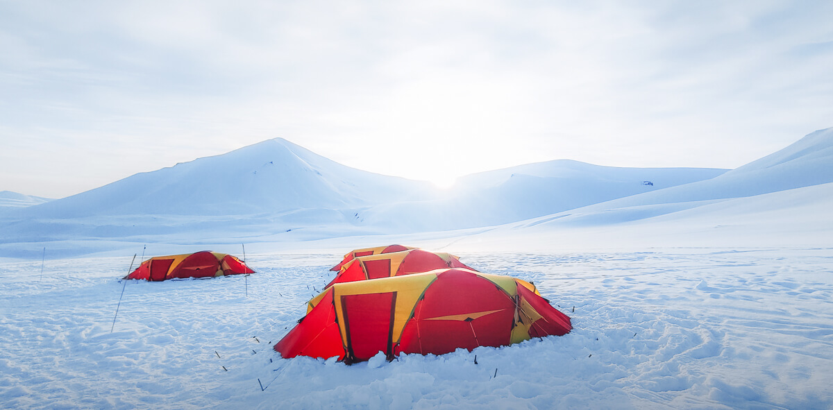 Red wild camping tents in a snowy Arctic landscape with distant mountains and blue sky.