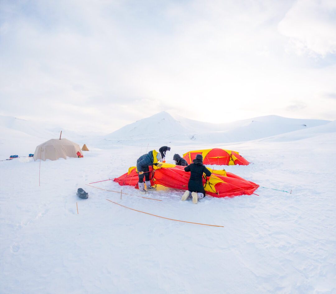 Women pitching a red snow tent and camping gear on Arctic snow in Svalbard during snowshoeing expedition