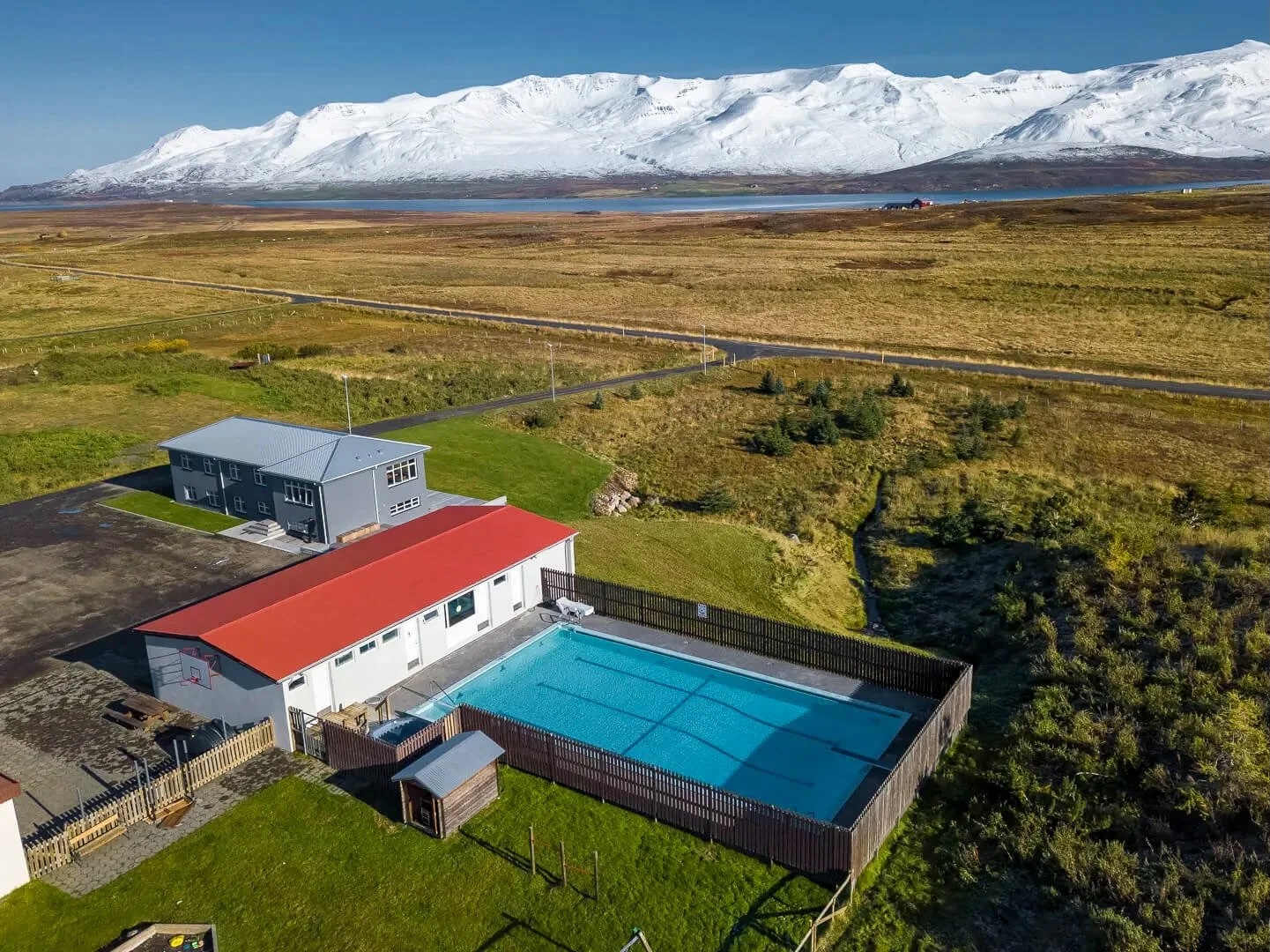 Aerial view of a lodge with outdoor swimming pool in open countryside beneath snow-covered mountains