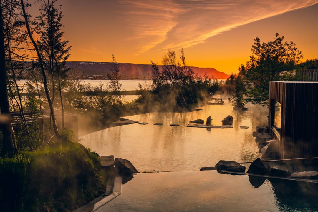Geothermal hot pool with steam and calm water at sunset by a river, with wooden fencing and trees nearby