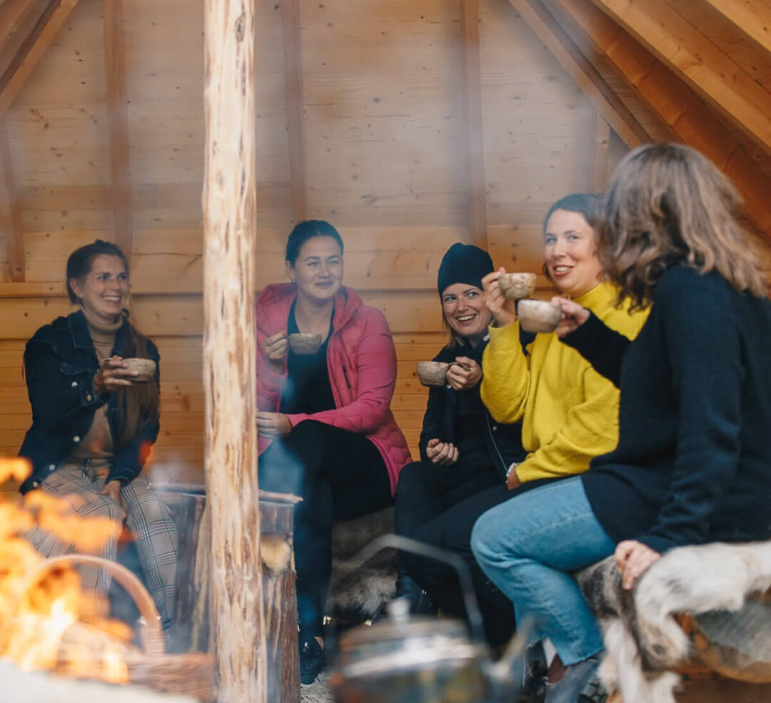 Women chatting and holding mugs by a fire inside a wooden lodge during a wilderness adventure trip