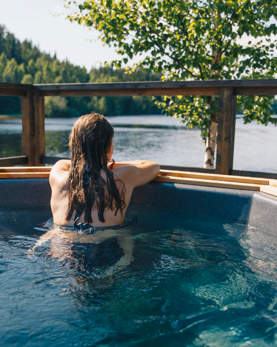 Woman relaxing in an outdoor hot tub beside a forest river, Swedish Lapland wilderness adventure