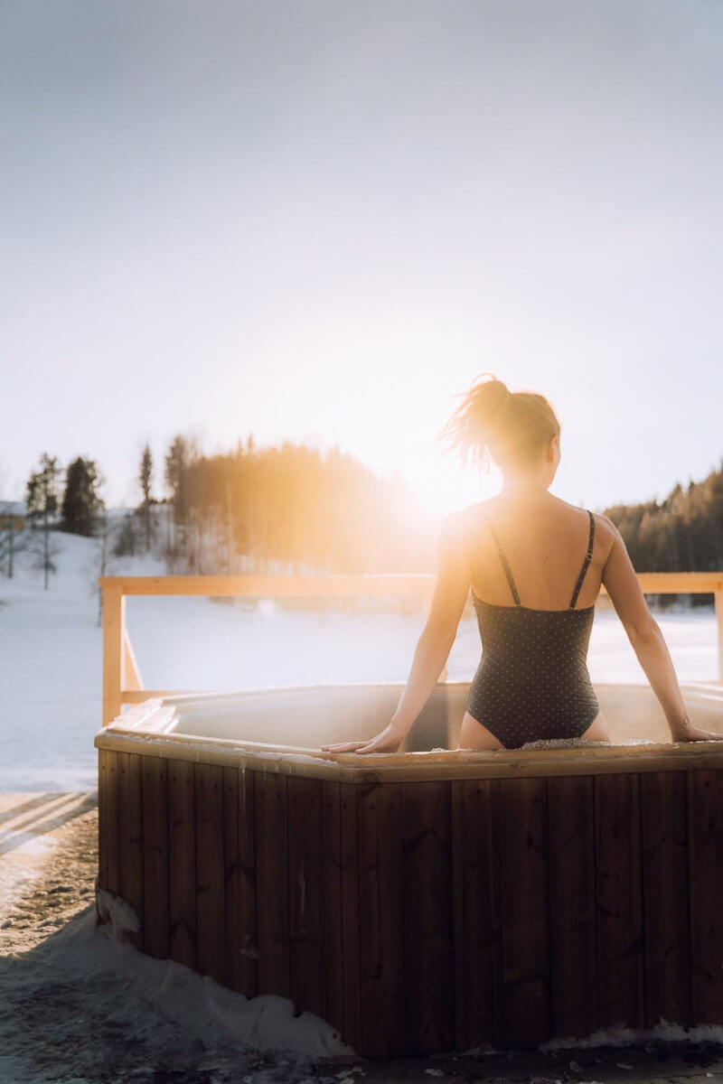Woman relaxing in an outdoor hot tub, steaming in a snowy forest at sunrise in Swedish Lapland