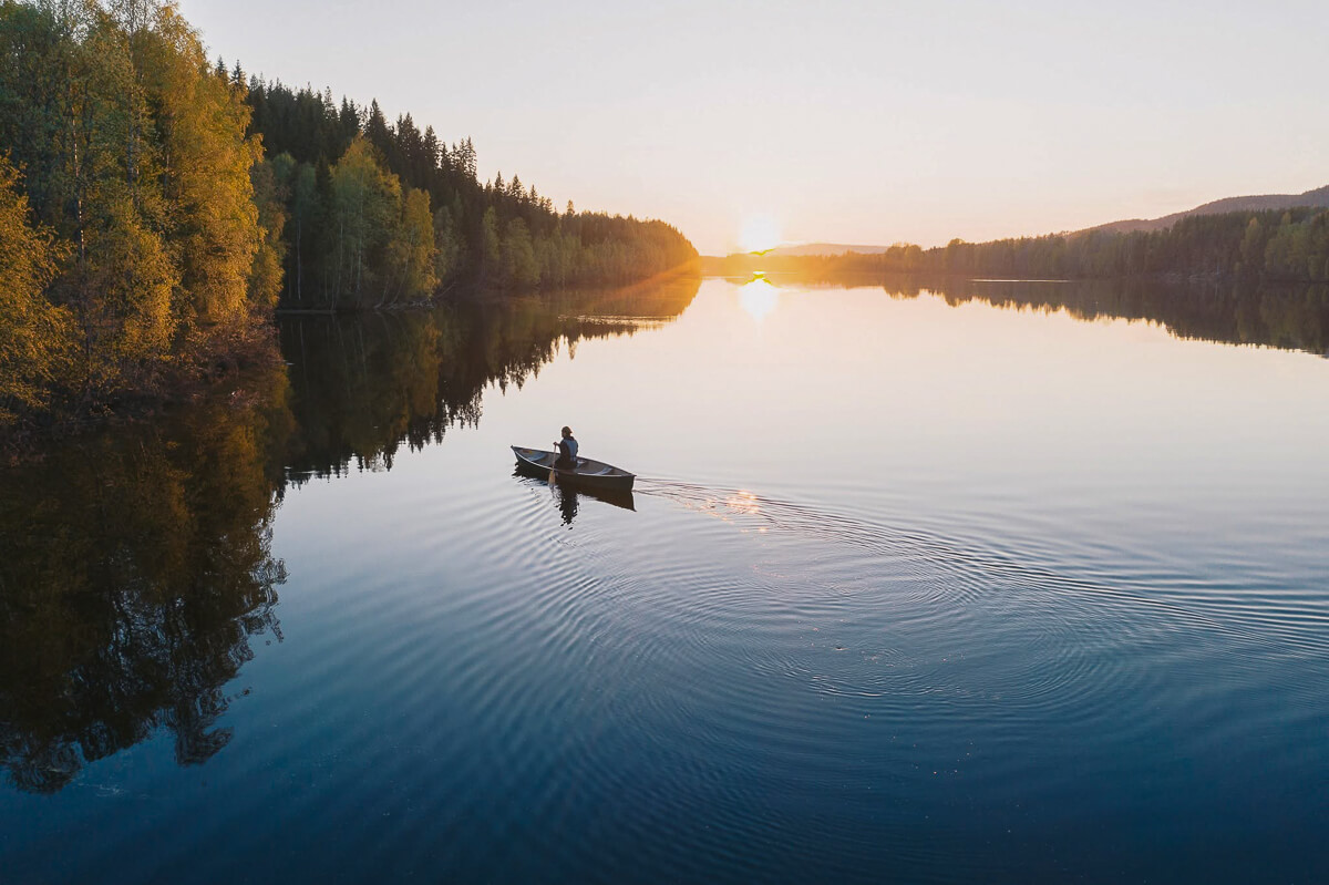 Woman in a small boat on a still lake at sunrise, surrounded by pine forests