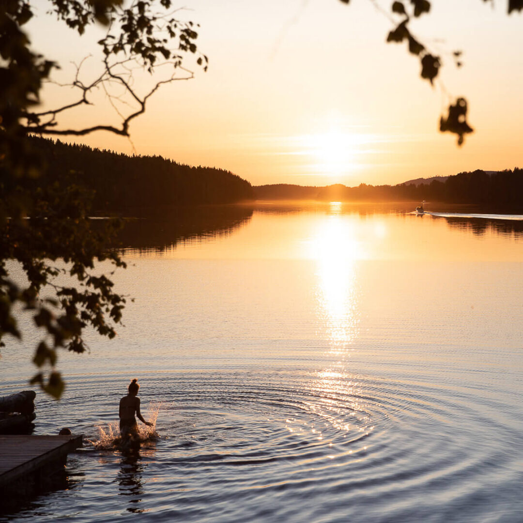 Woman wading in a calm lake at sunset, with forested shoreline and distant mountains