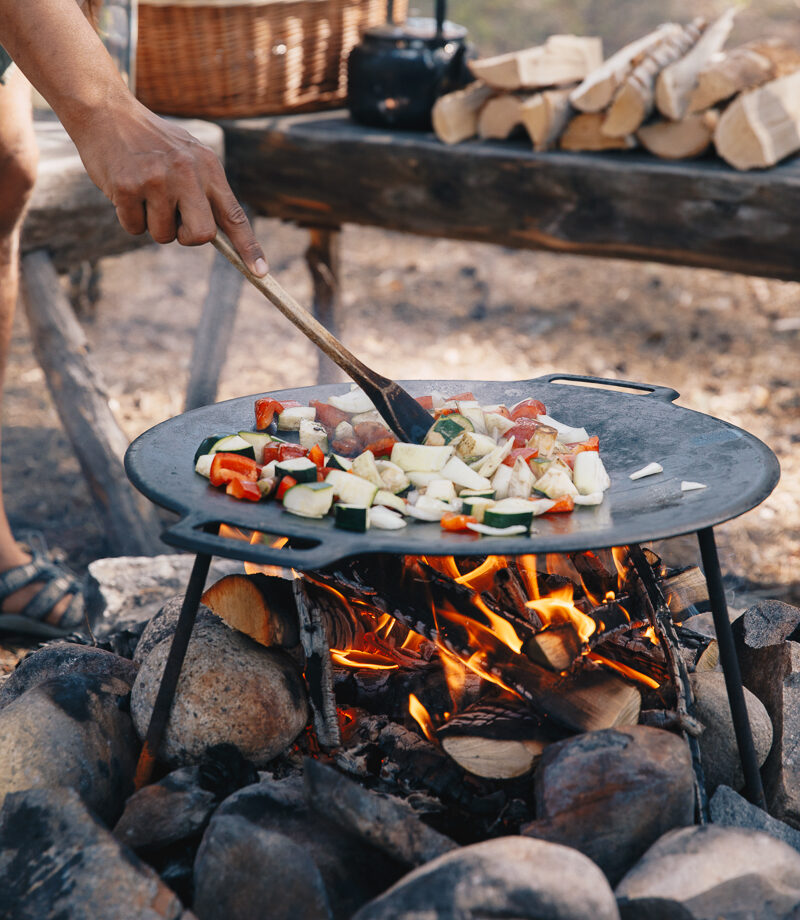 Cook stirring chopped vegetables in a frying pan over a campfire during an outdoor wilderness meal