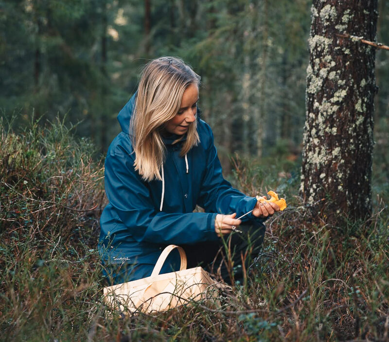 Woman foraging wild berries in a forest, wearing a blue jacket and carrying a white bag
