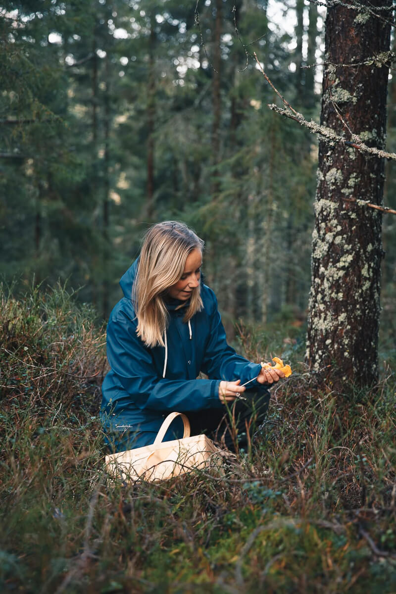 Woman in a blue jacket crouches in a forest, holding supplies and a bag during a wilderness adventure.