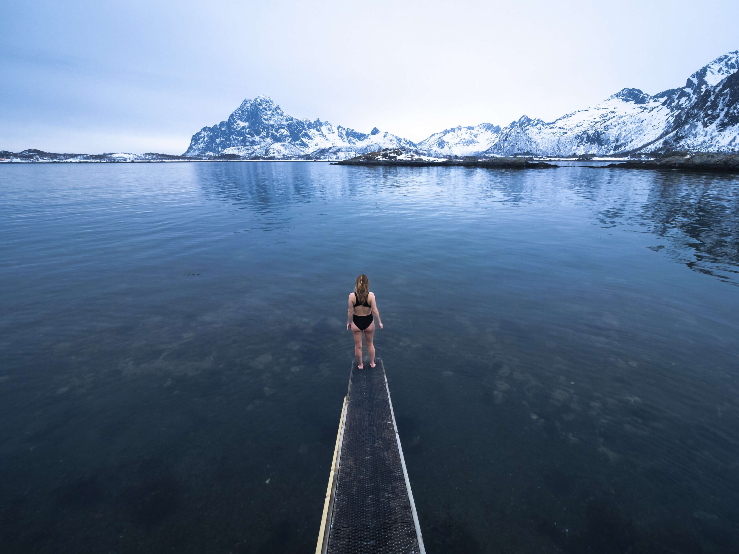 Woman standing on a kayak in a calm fjord with snowy Lofoten mountains in the background