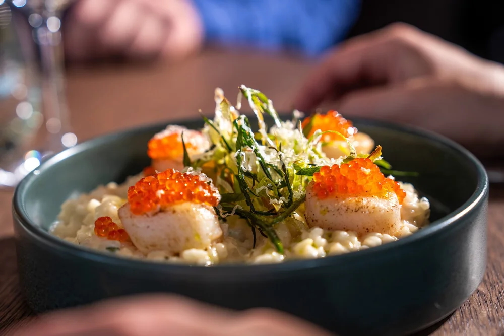 Seafood starter in a bowl with orange roe and greens, served at a table by two hands
