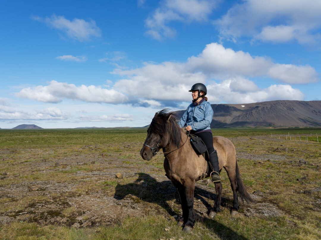 Woman horseback riding in Iceland, riding a brown horse across grassy landscape under a blue sky