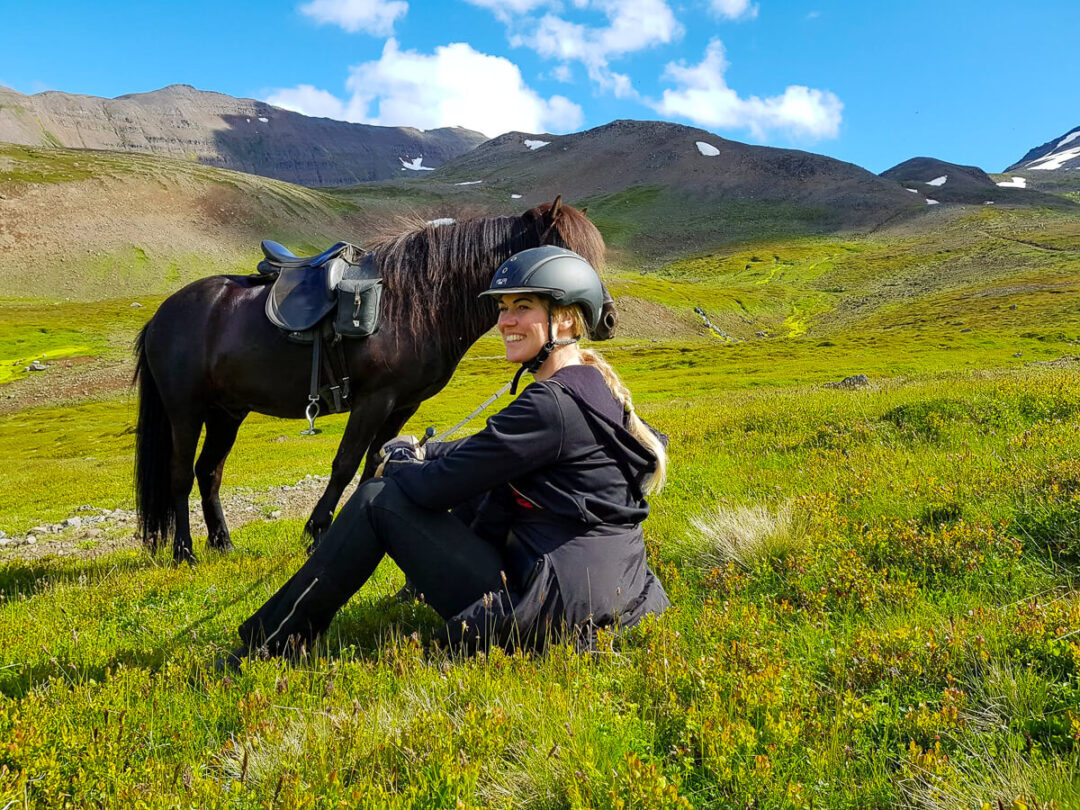 Woman wearing a helmet kneels beside a saddled horse in a grassy mountain valley outdoors