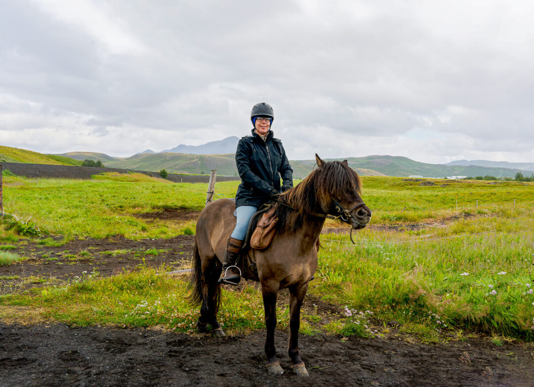 Woman riding a horse in Icelandic countryside, outdoors on a grassy trail with mountains in the distance
