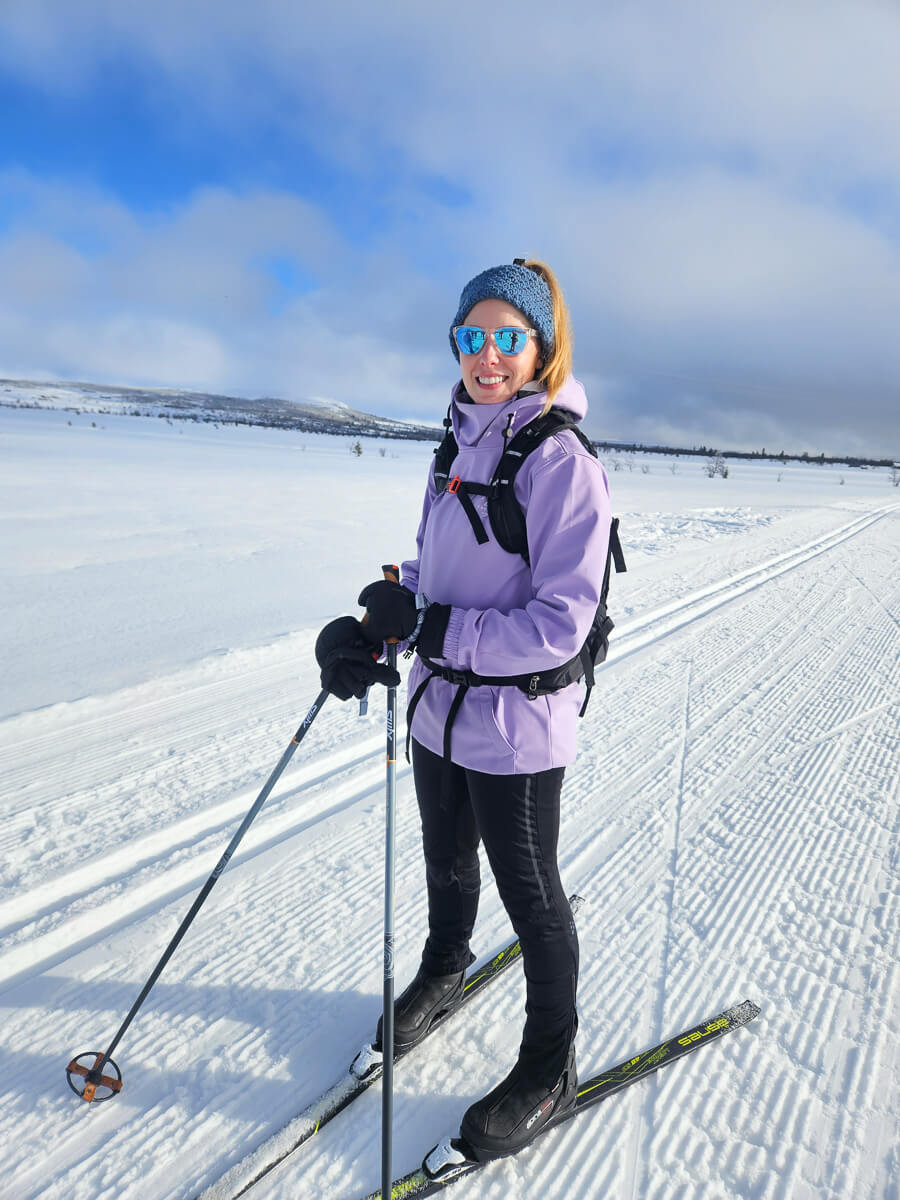 Woman cross-country skiing on snowy trail in open winter landscape, blue sky and tracks behind her