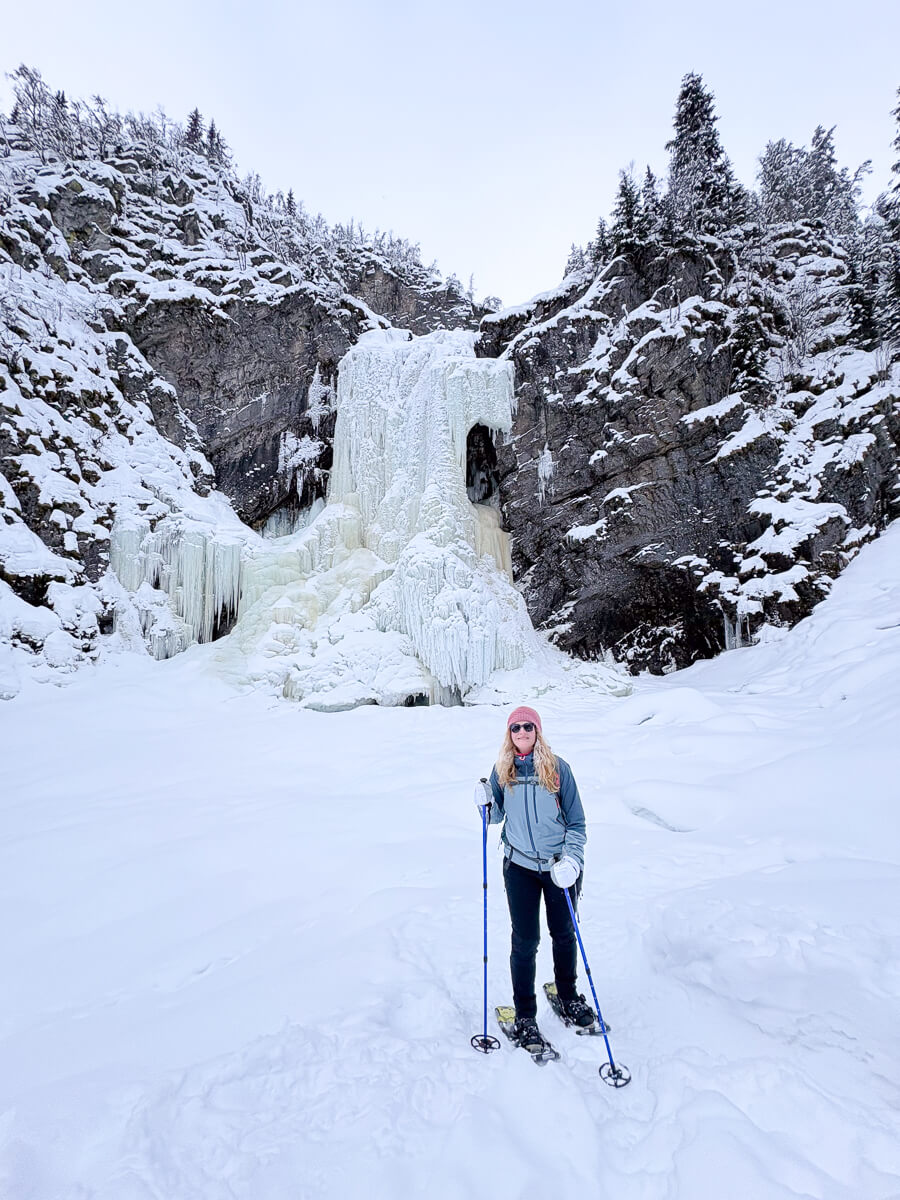 Woman cross-country skiing on a snowy mountain trail beside a frozen waterfall and fir trees