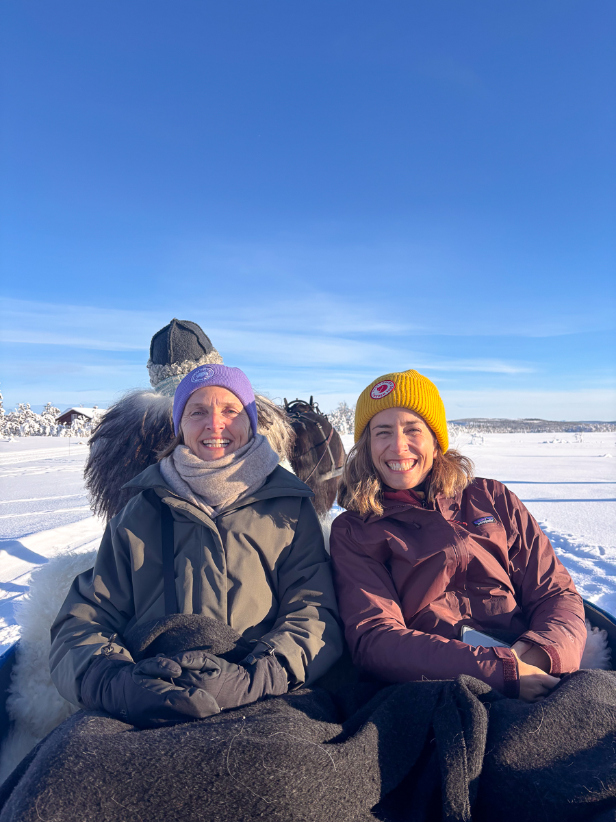Two women smiling in winter coats and hats on a snowy landscape in Norway.