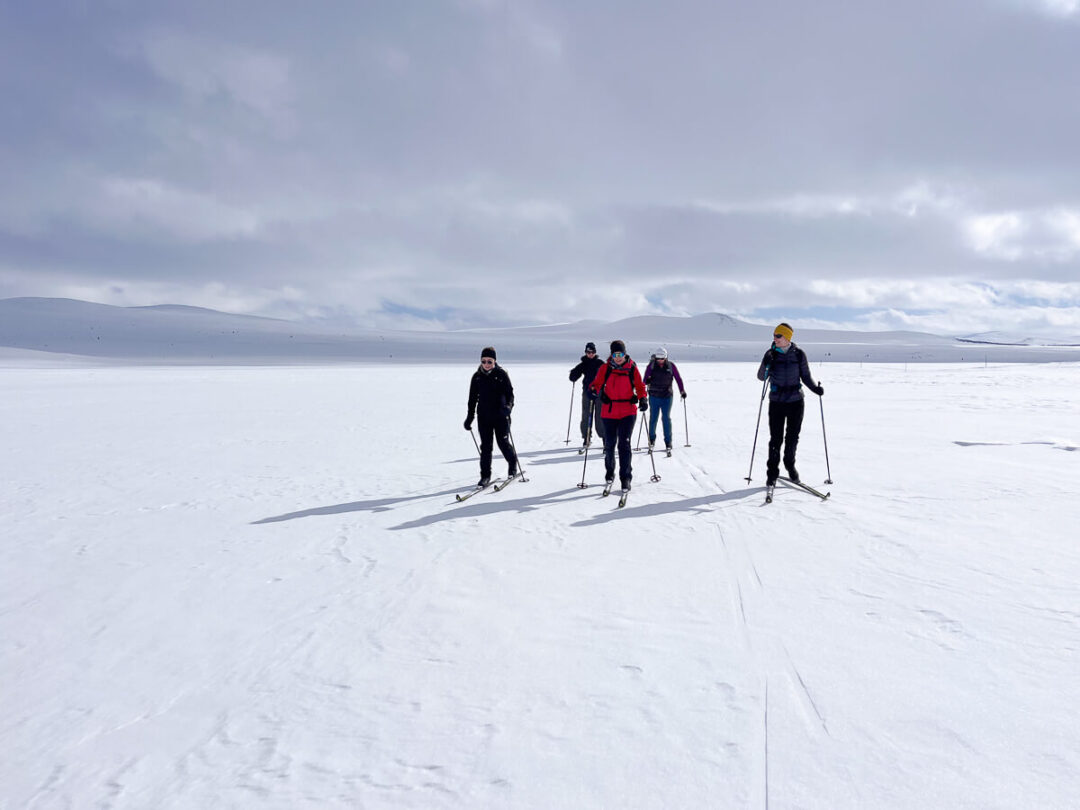 Women cross-country skiing across snowy Norwegian landscape in a group, with ski poles on a track.