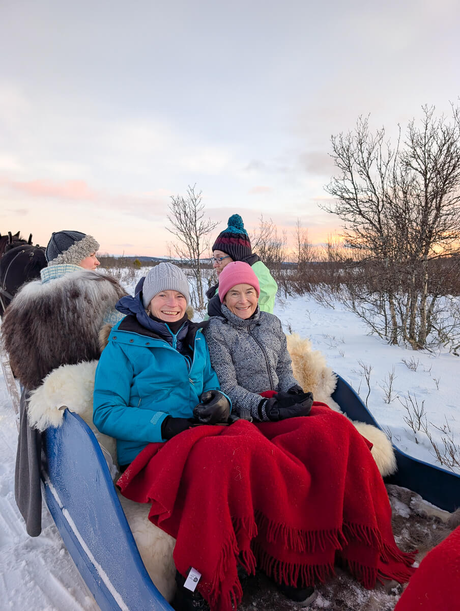 Women bundled up in winter coats on a horse-drawn sleigh in a snowy landscape at dusk