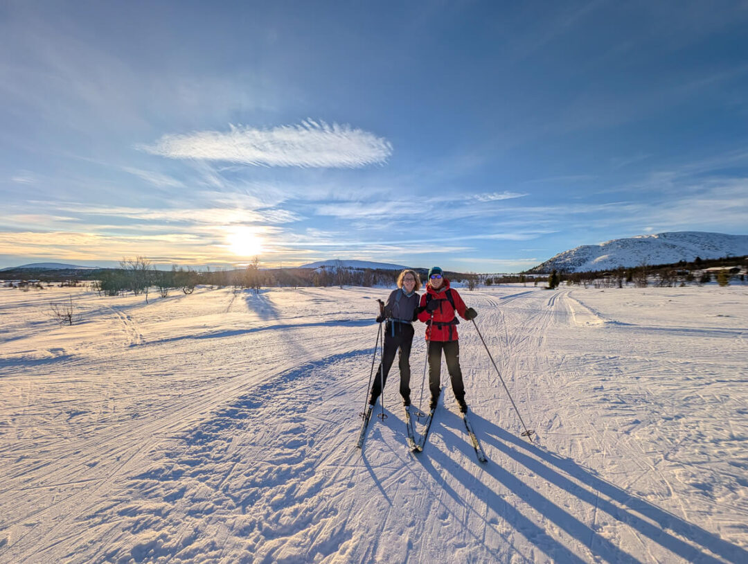 Two women cross-country skiing on a snowy trail at sunrise across open winter countryside