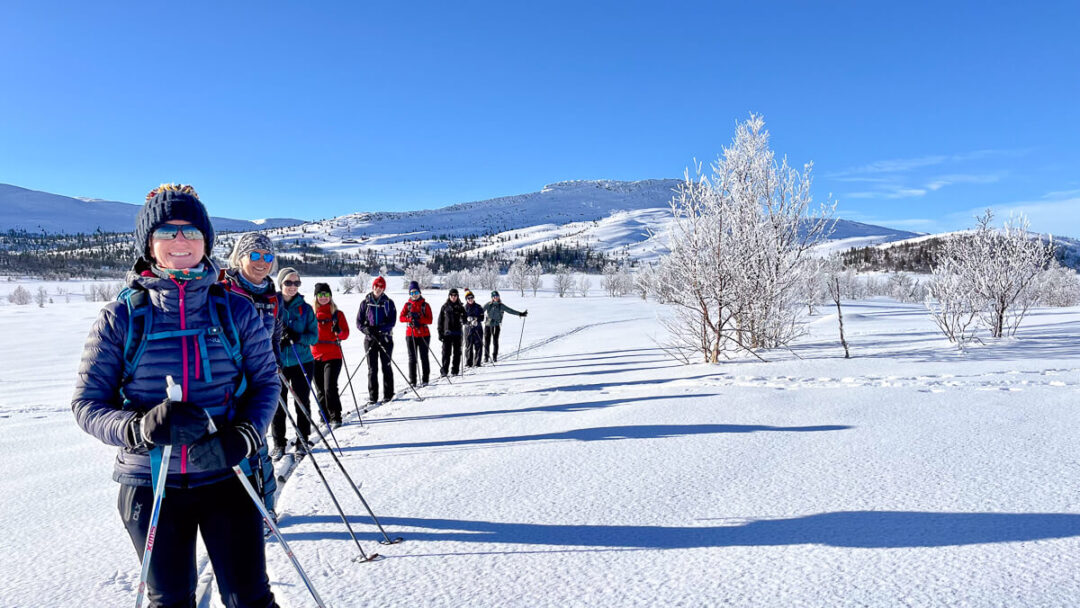 Women cross-country skiing on a snowy trail in Norway under blue sky, with a group behind