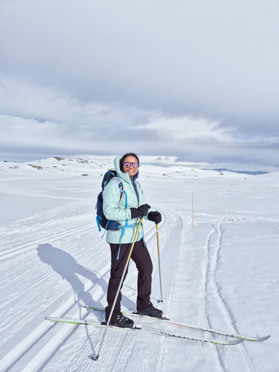 Woman beginner skier standing on a snowy ski slope with ski poles, under a cloudy sky.