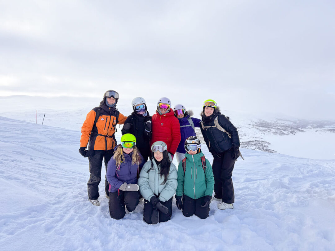 Women in ski helmets and jackets posing on a snowy mountain slope during skiing training.