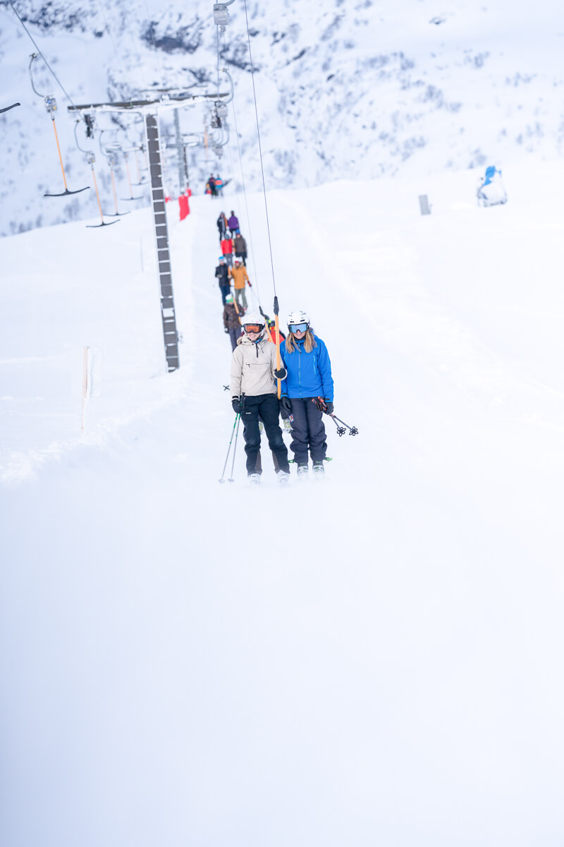 Women beginners on a ski chairlift in snowy mountains, wearing ski gear and holding skis