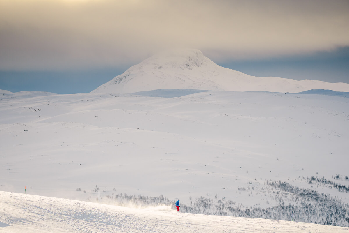 Woman skiing down a snowy mountain slope with a high peak in the distance under a cloudy sky